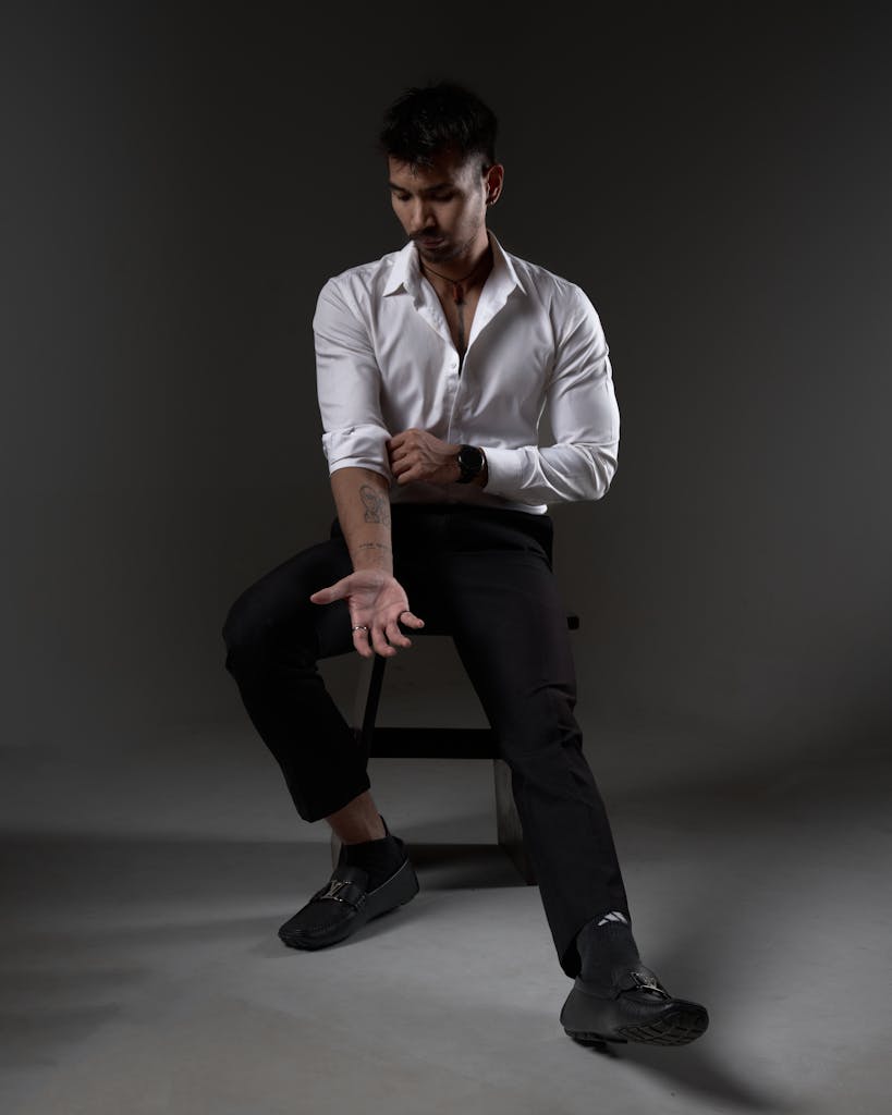 Portrait of a fashionable man in black pants and white shirt, sitting on a stool with an introspective expression.